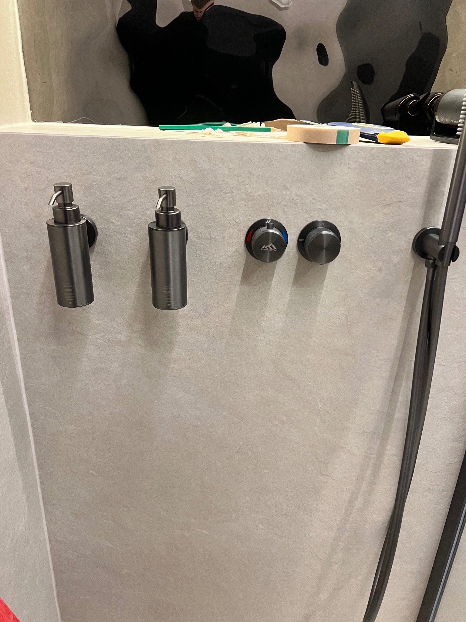 Nowoczesna łazienka: szare płytki, minimalistyczna armatura w kolorze grafitu, w tym dozowniki mydła i bateria prysznicowa z widocznym termostatem.