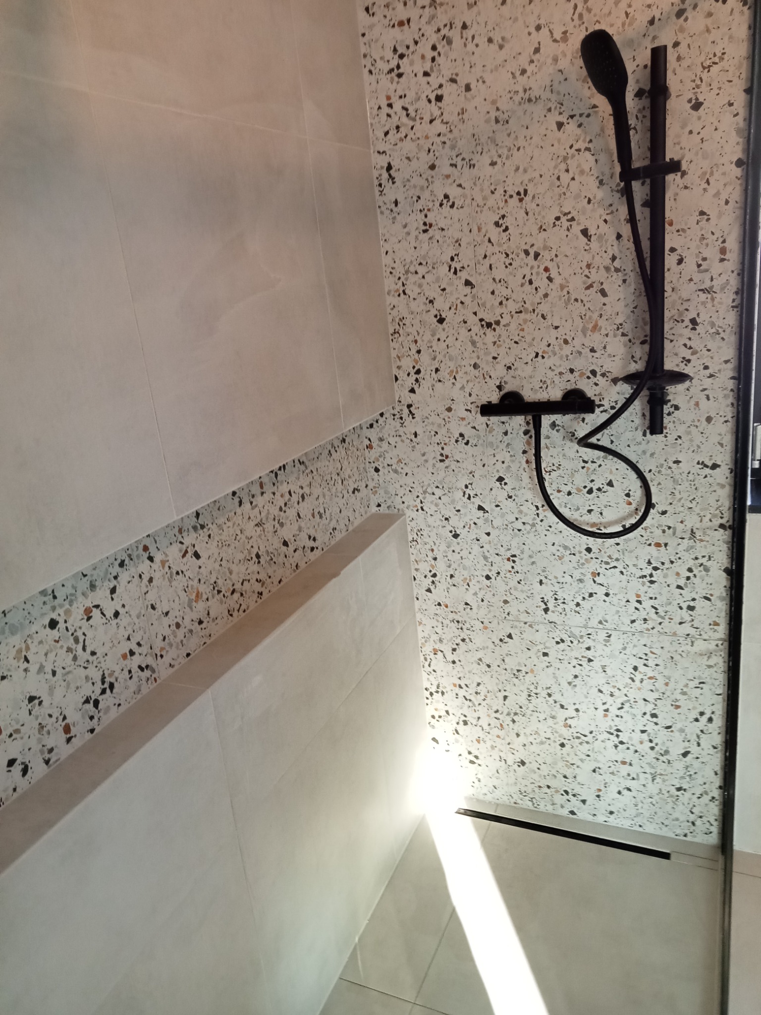 Nowoczesna kabina prysznicowa z czarną armaturą i ścianami wykończonymi płytkami imitującymi lastryko, połączonymi z jasnymi, gładkimi płytkami.