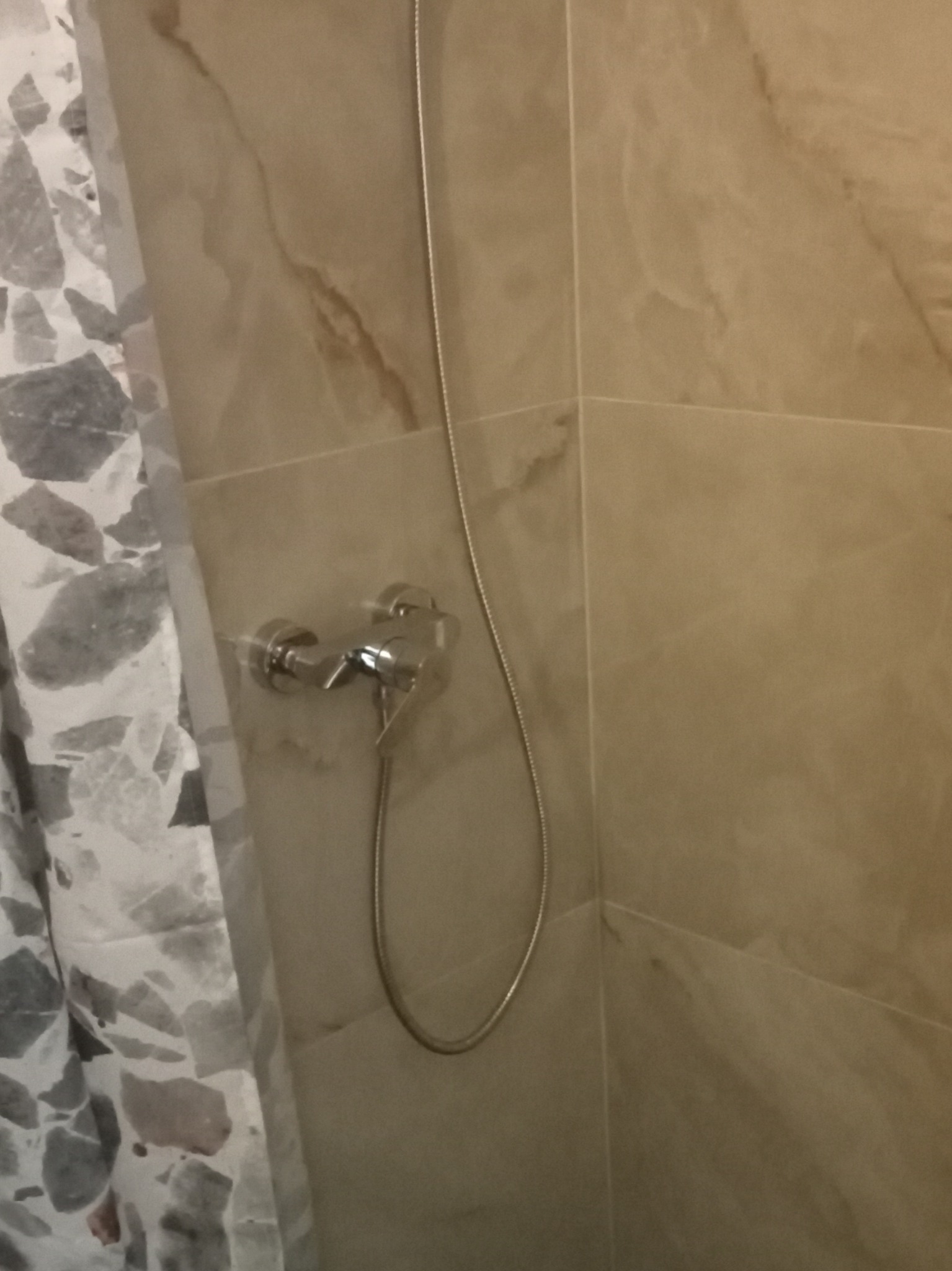 Beżowe płytki imitujące marmur w kabinie prysznicowej z chromowaną baterią i szarą zasłoną w geometryczne wzory.