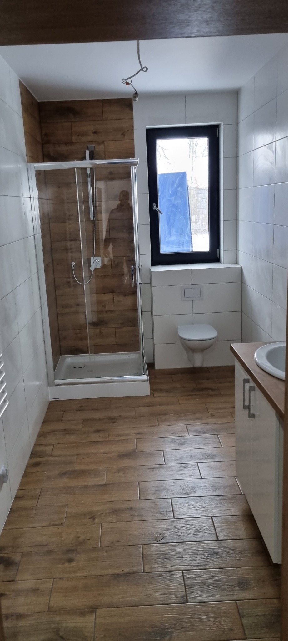 Nowoczesna łazienka z kabiną prysznicową ze szklanymi drzwiami, toaletą podwieszaną i umywalką z szafką, wykończona płytkami imitującymi drewno na podłodze i części ścian.