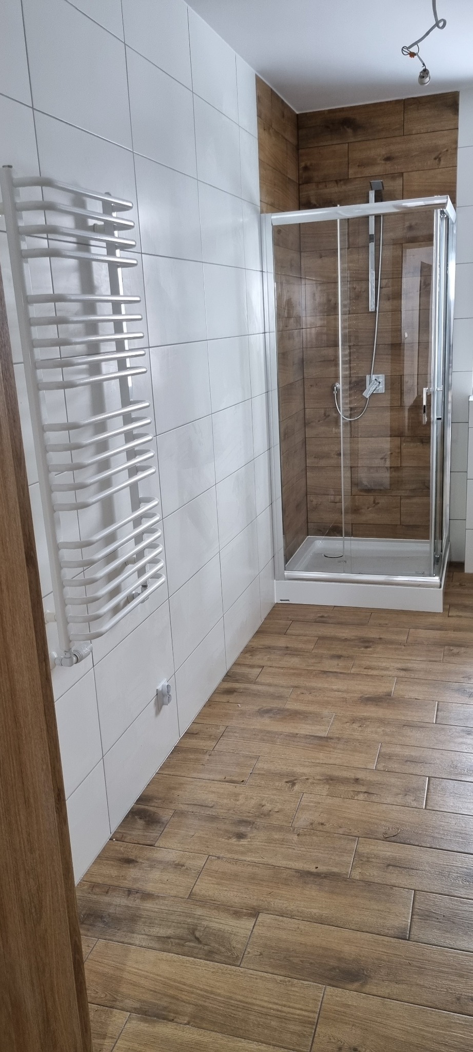 Nowoczesna łazienka z kabiną prysznicową wyłożoną płytkami imitującymi drewno, białe płytki na ścianach i grzejnik drabinkowy.