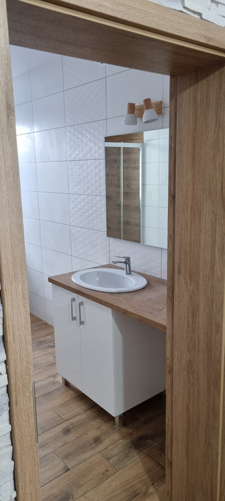 Nowoczesna łazienka z białą umywalką nablatową na szafce z drewnianym blatem, białe płytki z subtelnym wzorem na ścianie i drewnopodobna podłoga, widoczna kabina prysznicowa w lustrze.