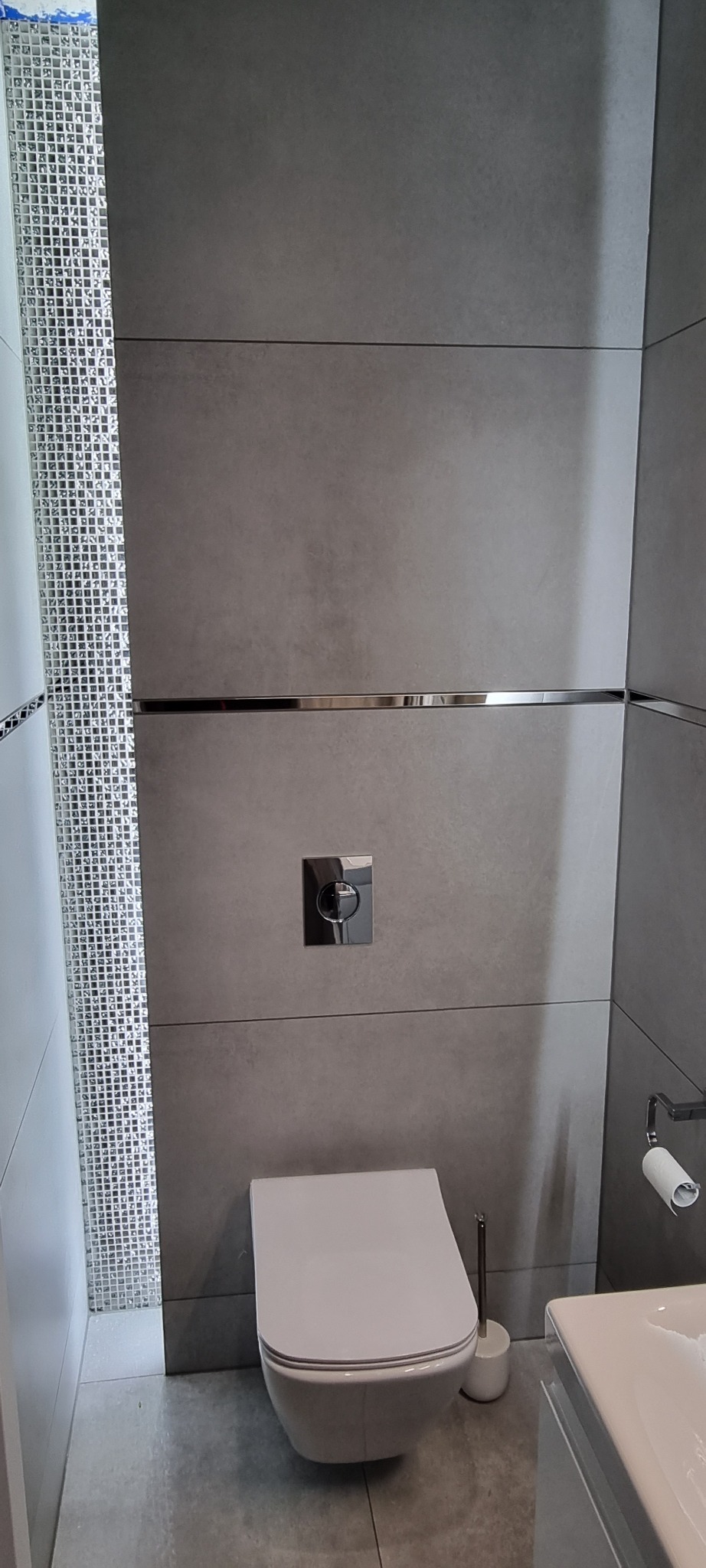 Nowoczesna toaleta podwieszana z szarymi płytkami, dekoracyjną mozaiką i chromowanymi listwami w wykończonej łazience.