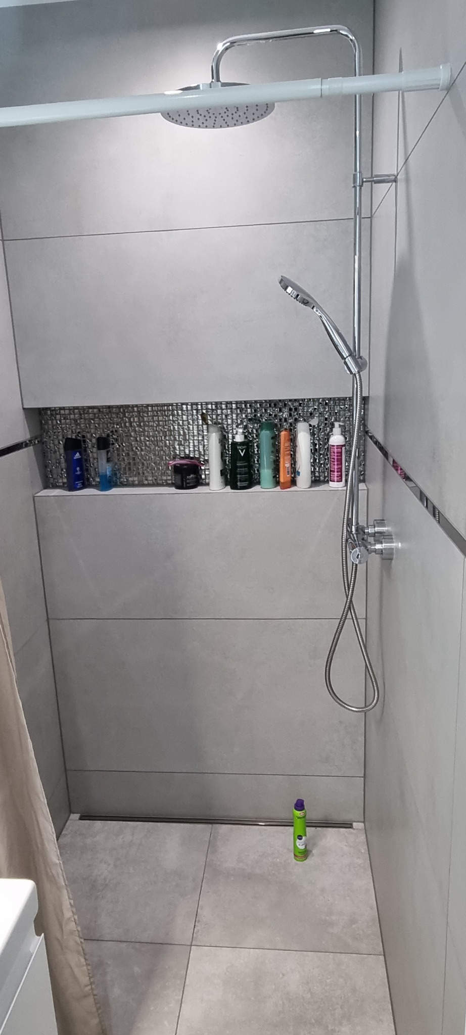 Nowoczesna kabina prysznicowa z szarymi płytkami, deszczownicą i półką z kosmetykami, widoczny odpływ liniowy i butelka środka czyszczącego na podłodze.