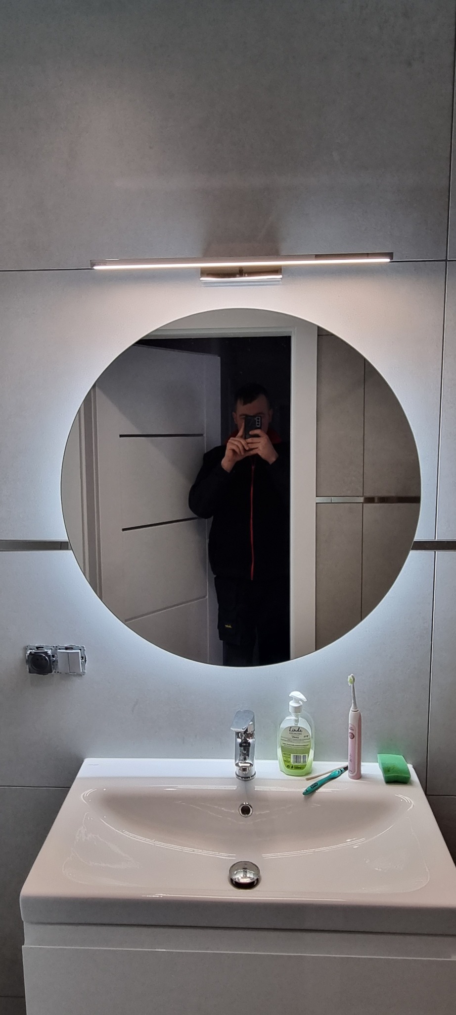 Nowoczesna łazienka z okrągłym lustrem podświetlanym LED, szara płytka na ścianie, biała umywalka z baterią, mydło w płynie, szczoteczka do zębów i odbicie osoby robiącej zdjęcie.