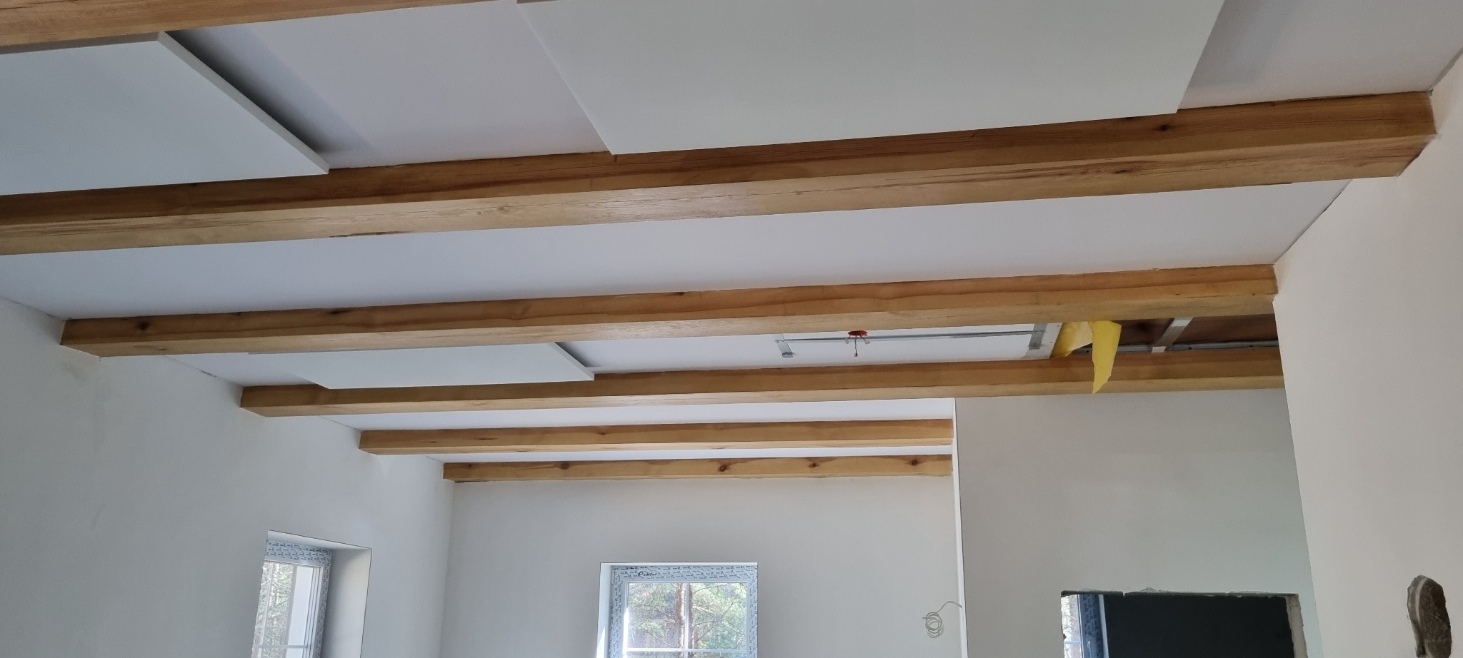 Poddasze z widocznymi drewnianymi belkami stropowymi i częściowo zamontowanymi białymi panelami sufitowymi, z widoczną instalacją i ociepleniem z wełny mineralnej.