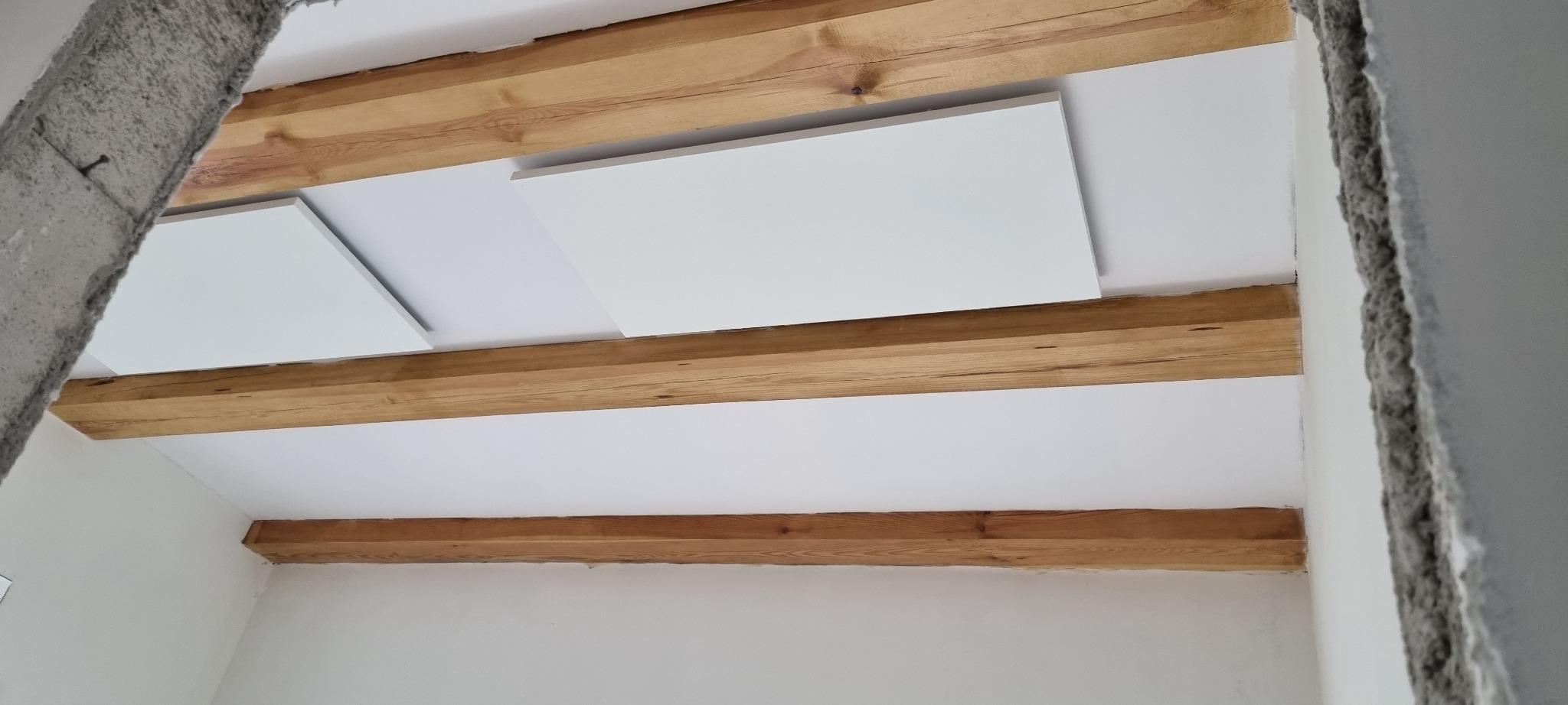 Surowy stan wykończenia wnętrza: drewniane belki stropowe, białe płyty sufitowe i fragment ściany w stanie deweloperskim.