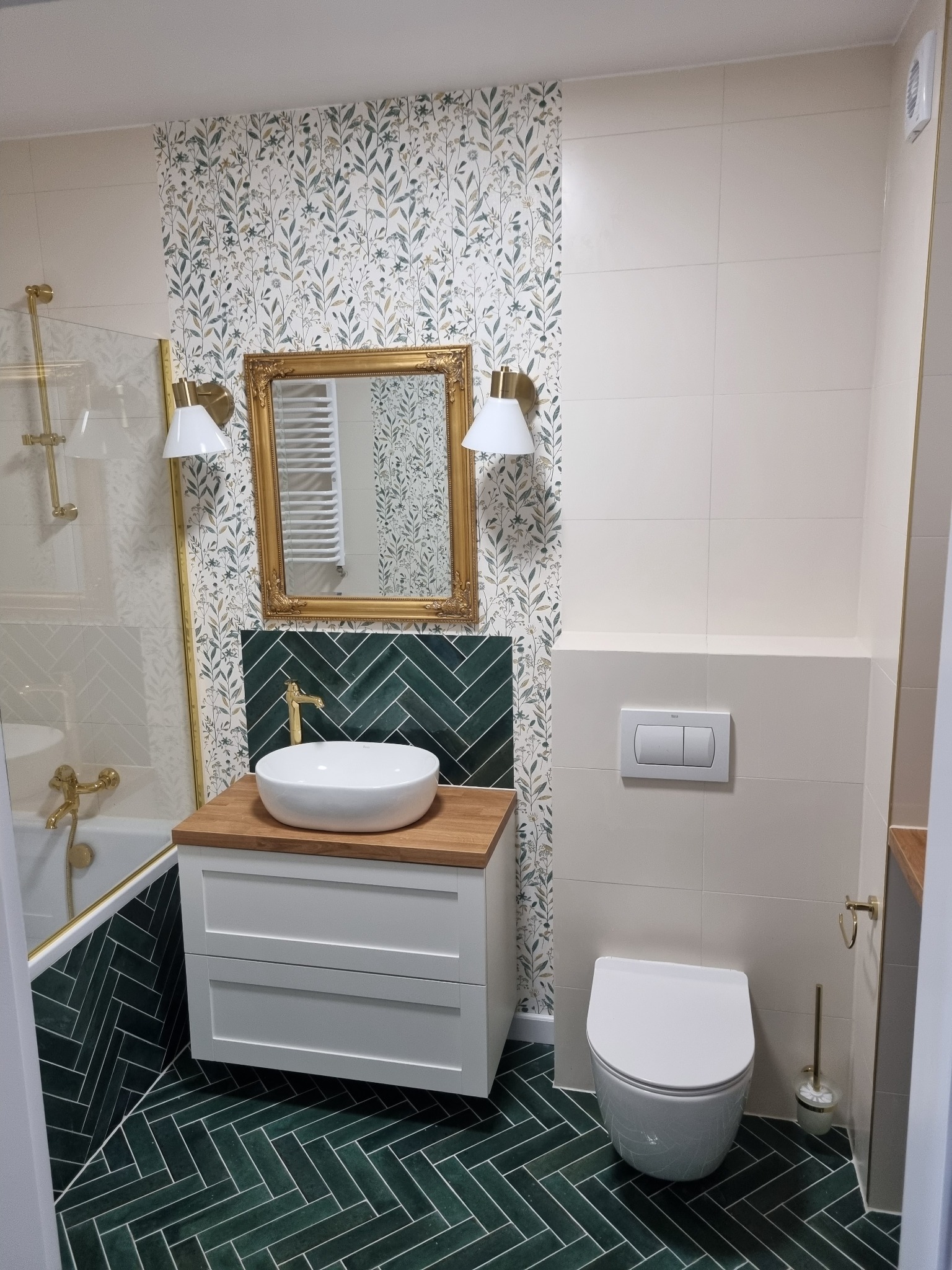 Zakończona aranżacja łazienki z zieloną, ułożoną w jodełkę glazurą na podłodze i ścianie, białą ceramiką sanitarną, drewnianym blatem pod umywalką i lustrem w złotej ramie.
