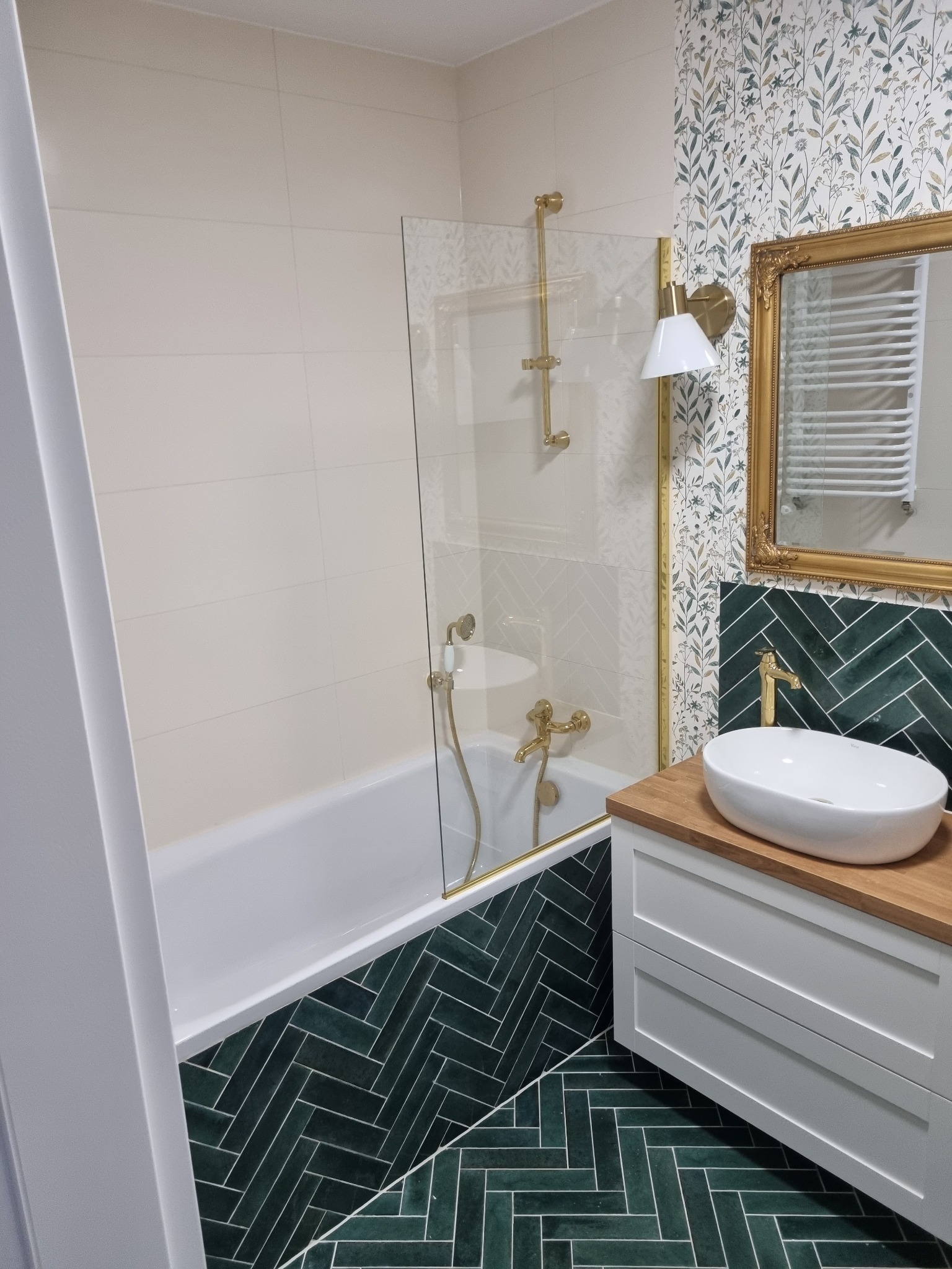 Nowoczesna łazienka z zielonymi płytkami ułożonymi w jodełkę, złotą armaturą, umywalką nablatową na drewnianym blacie i tapetą w roślinne wzory.
