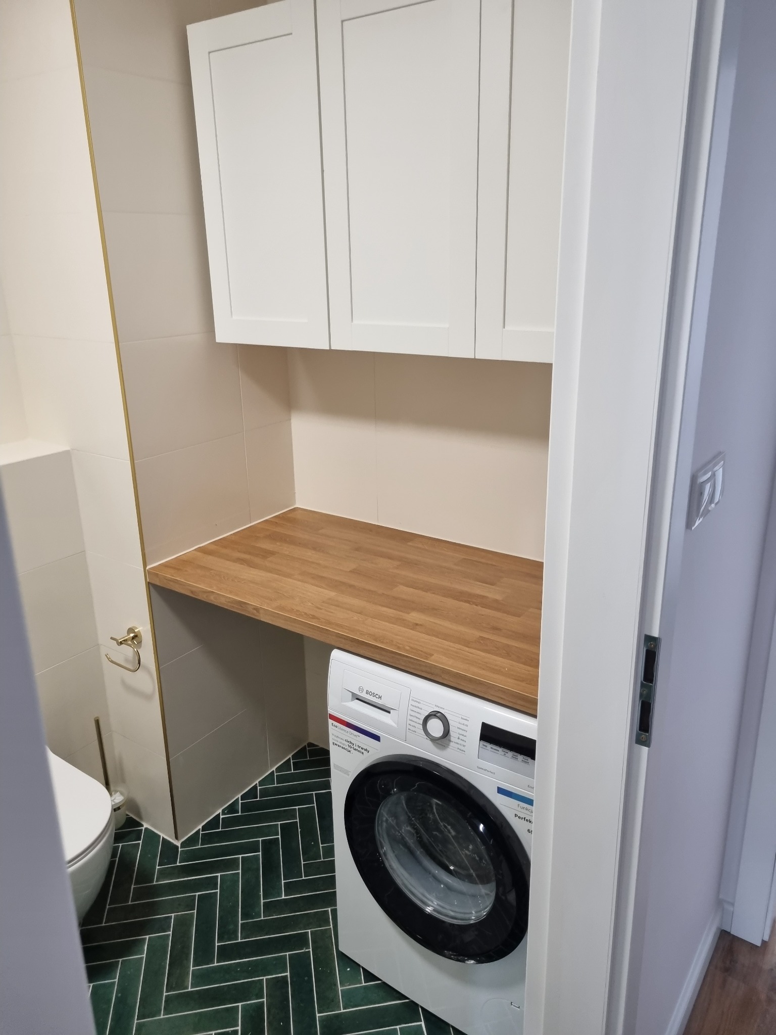 Wykończona łazienka w Dąbrowie Białostockiej z zieloną podłogą w jodełkę, białą pralką Bosch pod drewnianym blatem i szafkami wiszącymi.