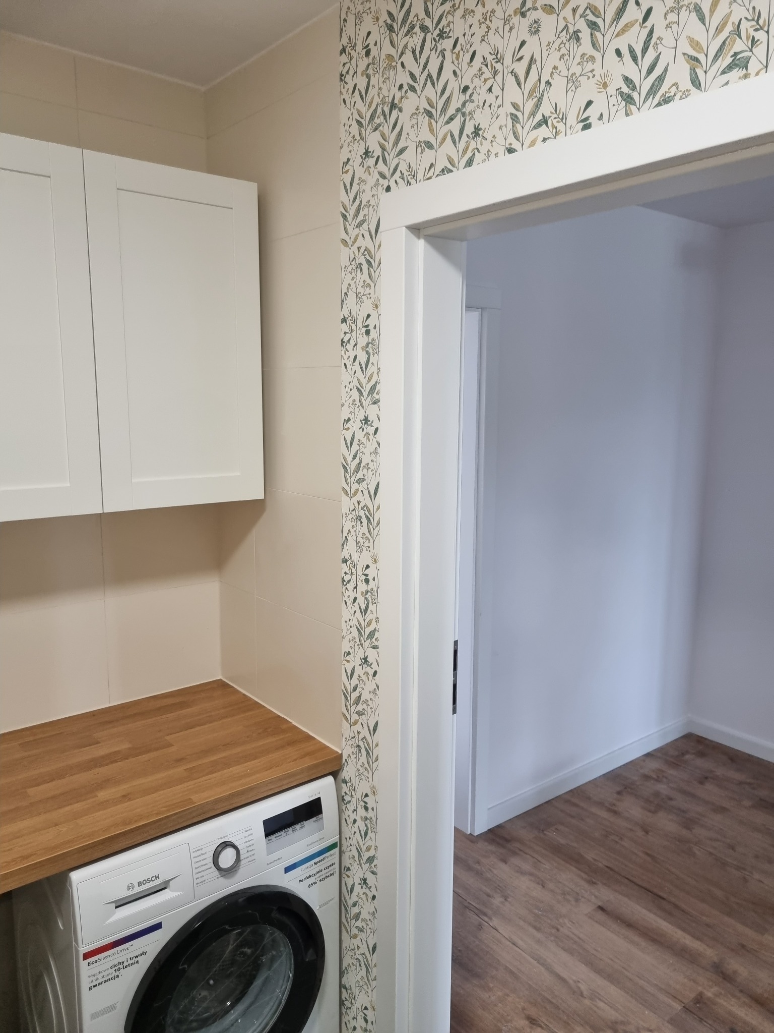 Nowoczesna łazienka z białą zabudową szafek nad pralką Bosch, drewnianym blatem, beżowymi płytkami i tapetą w roślinne motywy, widok na sąsiednie pomieszczenie z parkietem.