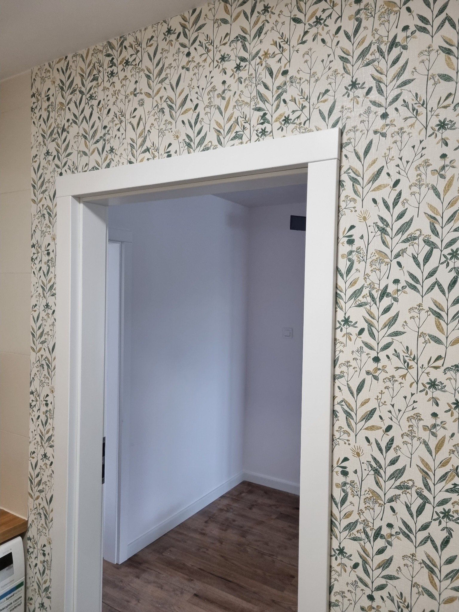 Ściana z tapetą w roślinny wzór, widok na białe futryny drzwi i podłogę z paneli w przejściu do pomieszczenia.