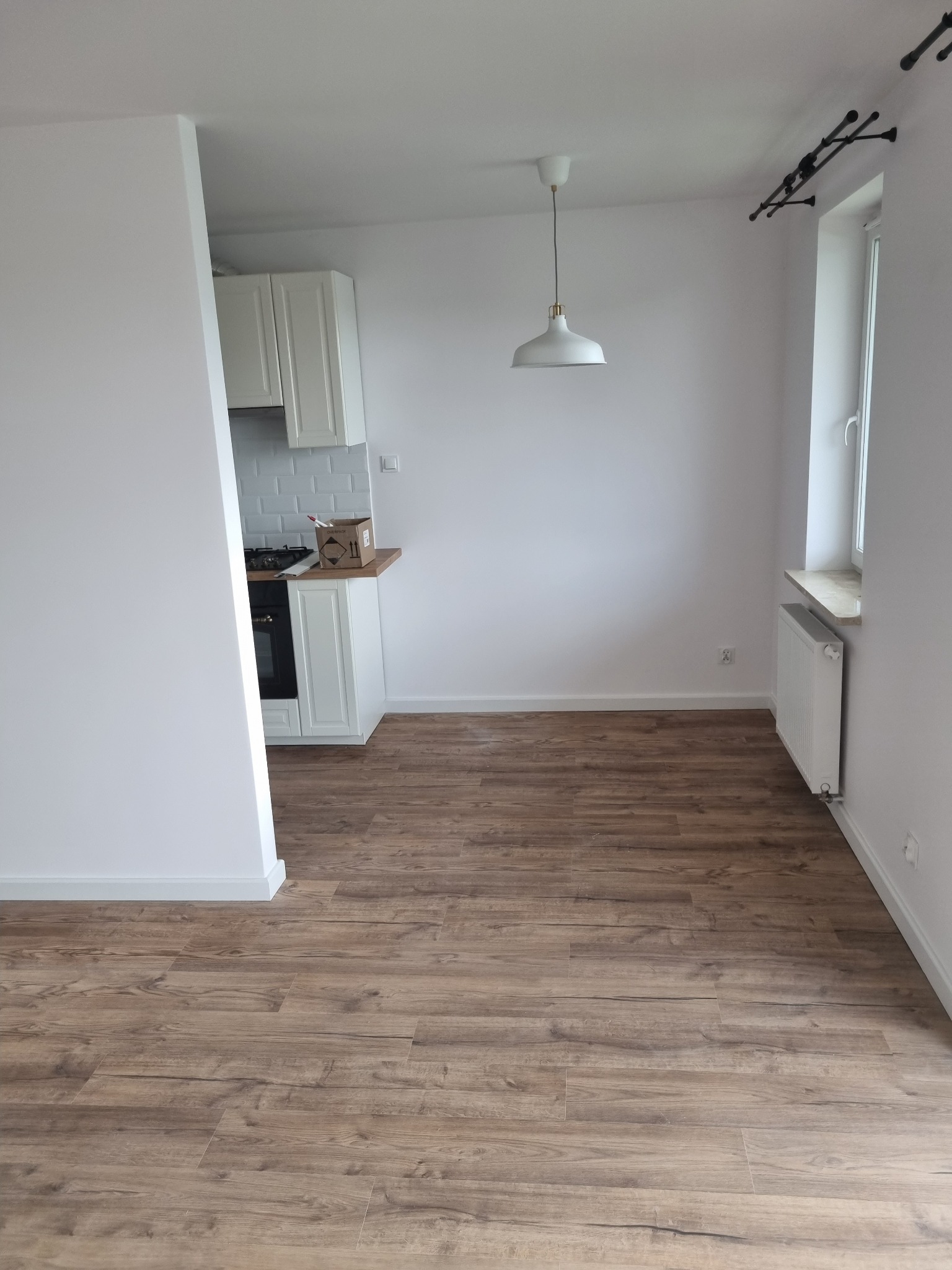 Puste, jasne wnętrze mieszkania po remoncie z drewnopodobną podłogą, białymi ścianami, grzejnikiem pod oknem i widocznym fragmentem zabudowy kuchennej z białą glazurą.