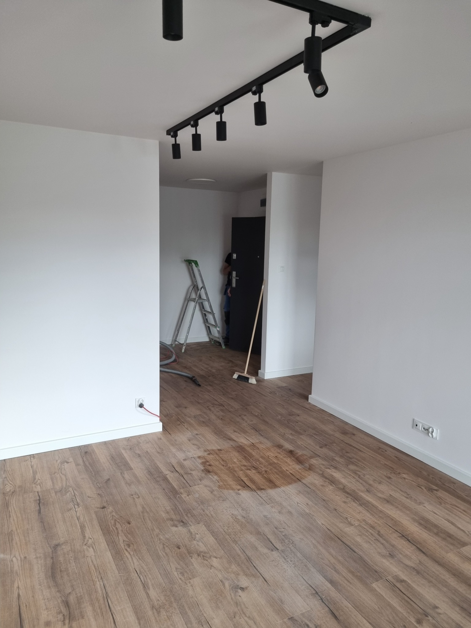 Pomalowane na biało wnętrze mieszkania z ułożonymi panelami podłogowymi, widoczna drabina, odkurzacz i lampa sufitowa na szynie.