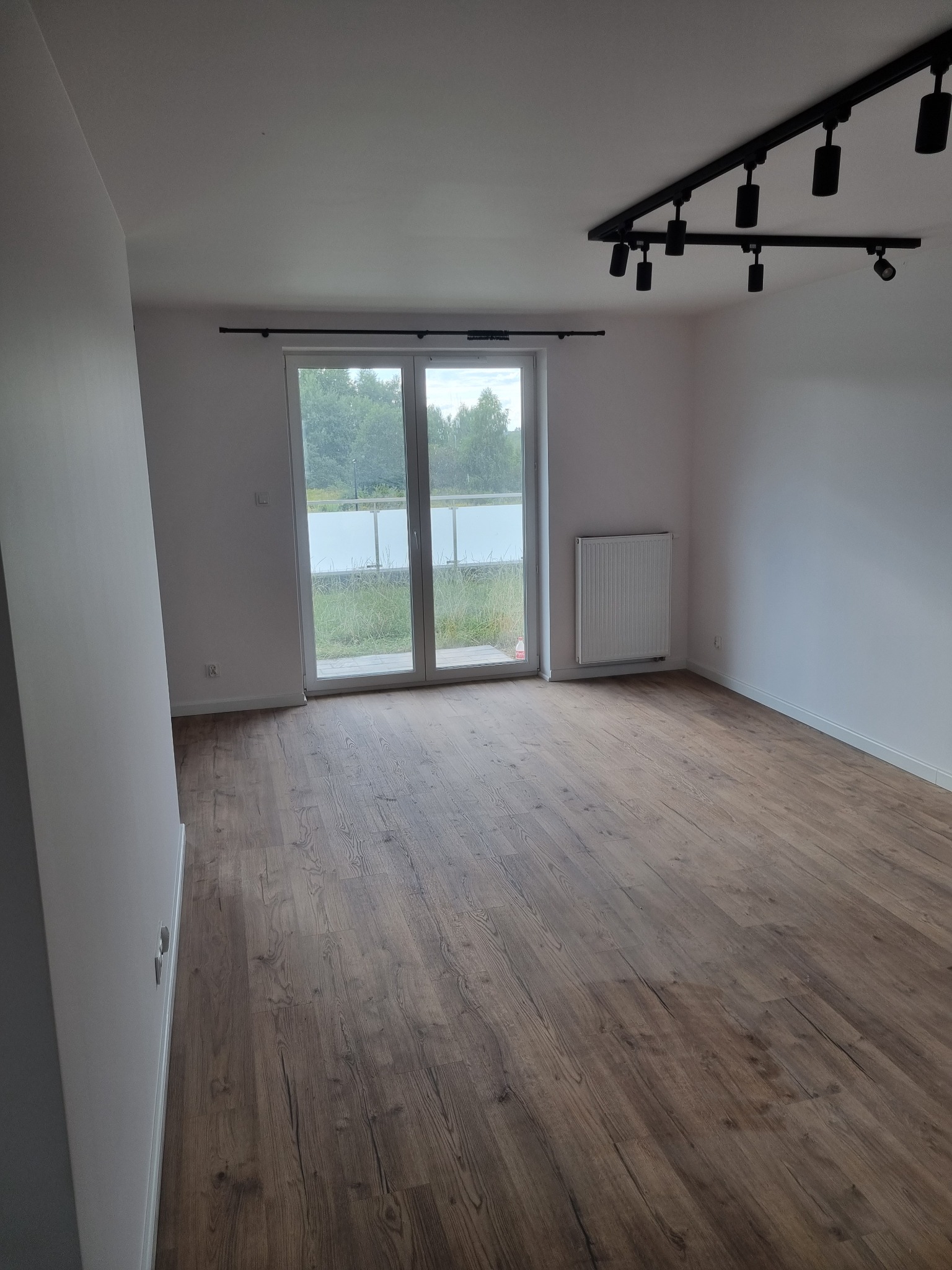 Pusty pokój z panelami podłogowymi w odcieniach brązu, białymi ścianami, grzejnikiem i oknem balkonowym z widokiem na zieleń. Na suficie czarna listwa z reflektorami.