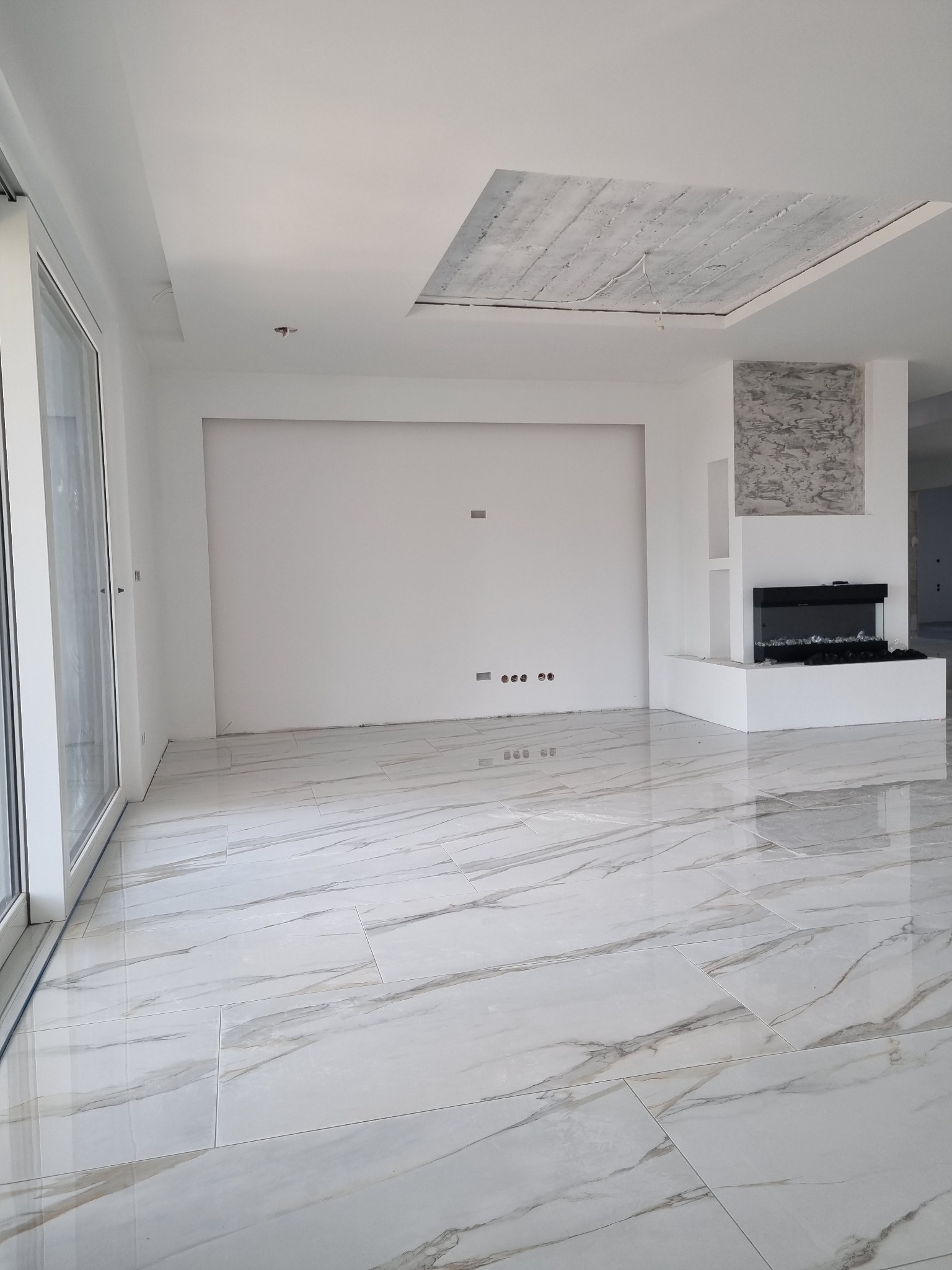 Pusty, jasny pokój z marmurową podłogą, elektrycznym kominkiem i surowym betonowym elementem na suficie, widoczne gniazdka elektryczne na ścianie.