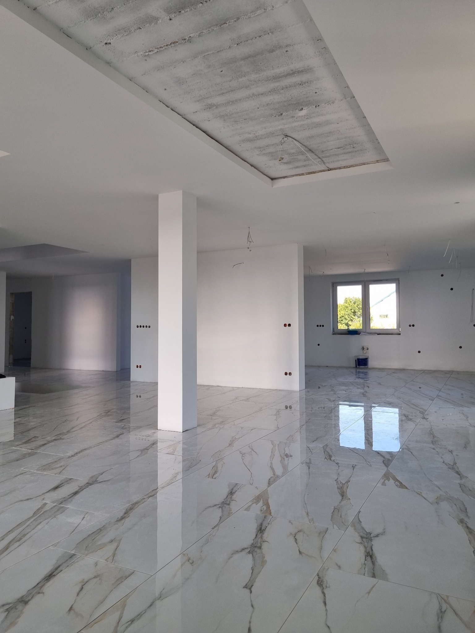 Wnętrze w stanie deweloperskim z błyszczącą, marmurkową podłogą, surowym betonowym fragmentem sufitu, białymi ścianami z widocznymi puszkami elektrycznymi i kolumną nośną.