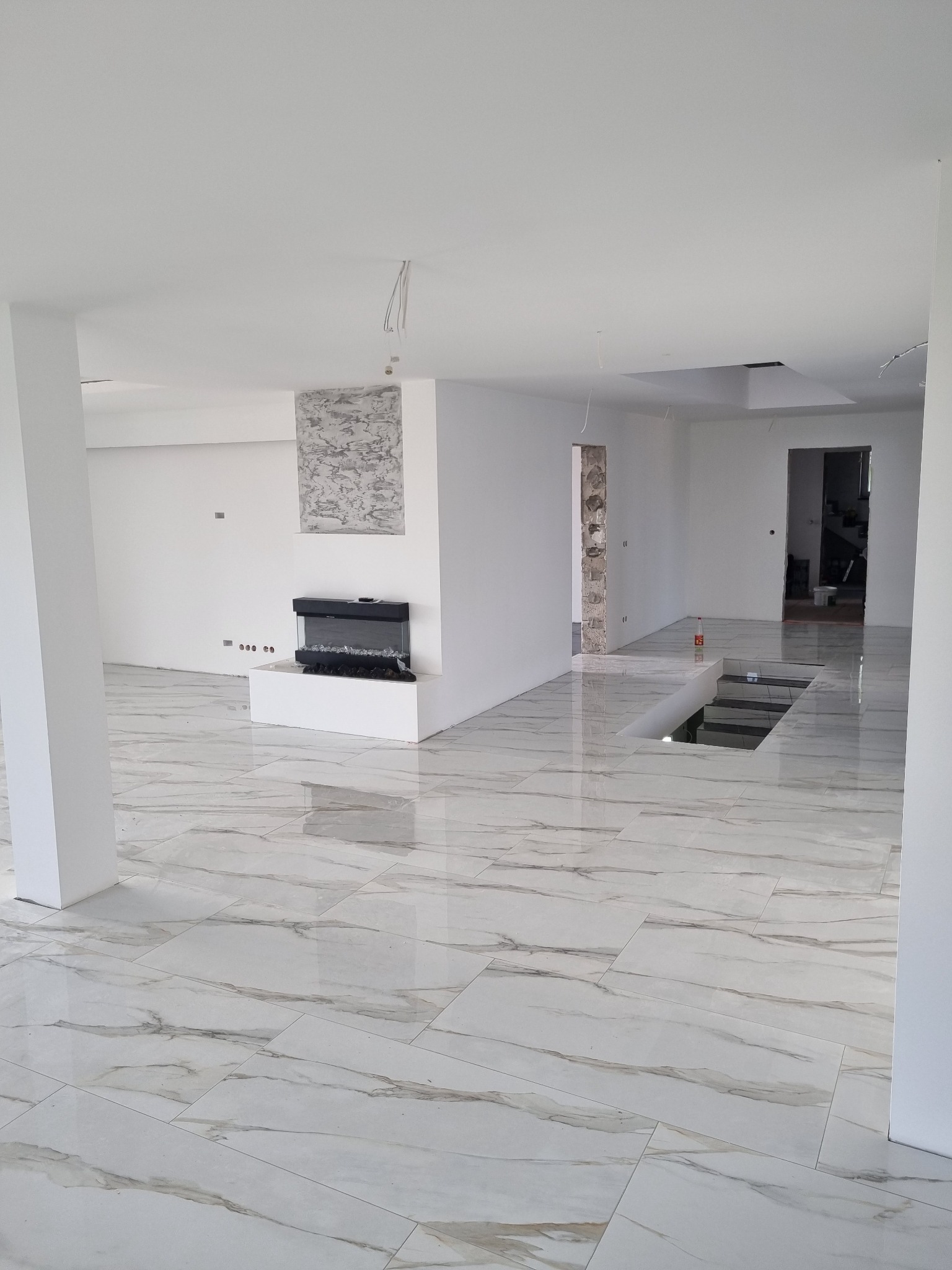 Przestronne, jasne wnętrze w trakcie remontu z błyszczącą, marmurkową podłogą, elektrycznym kominkiem i przygotowanym zejściem do piwnicy.