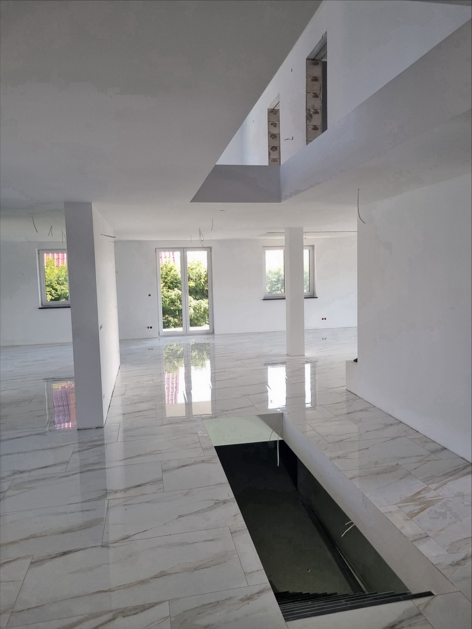 Przestronne wnętrze w stanie deweloperskim z błyszczącą podłogą imitującą marmur, widoczne filary i okna z zielenią na zewnątrz, w tle surowe ściany i schody prowadzące w dół.