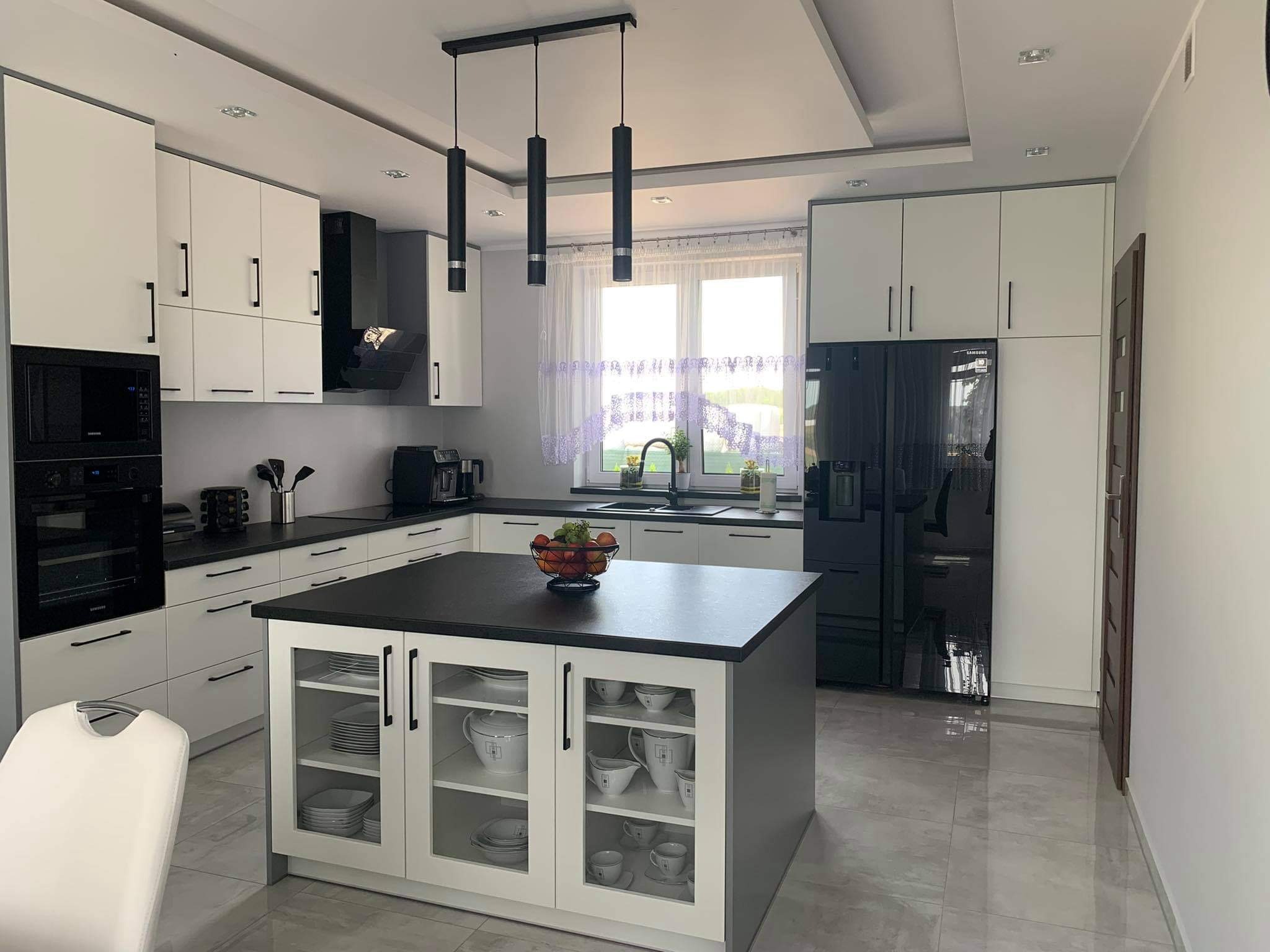 Nowoczesna kuchnia w bieli i czerni z wyspą, na której stoi kosz z owocami. Widoczne czarne, wiszące lampy i sprzęt AGD.