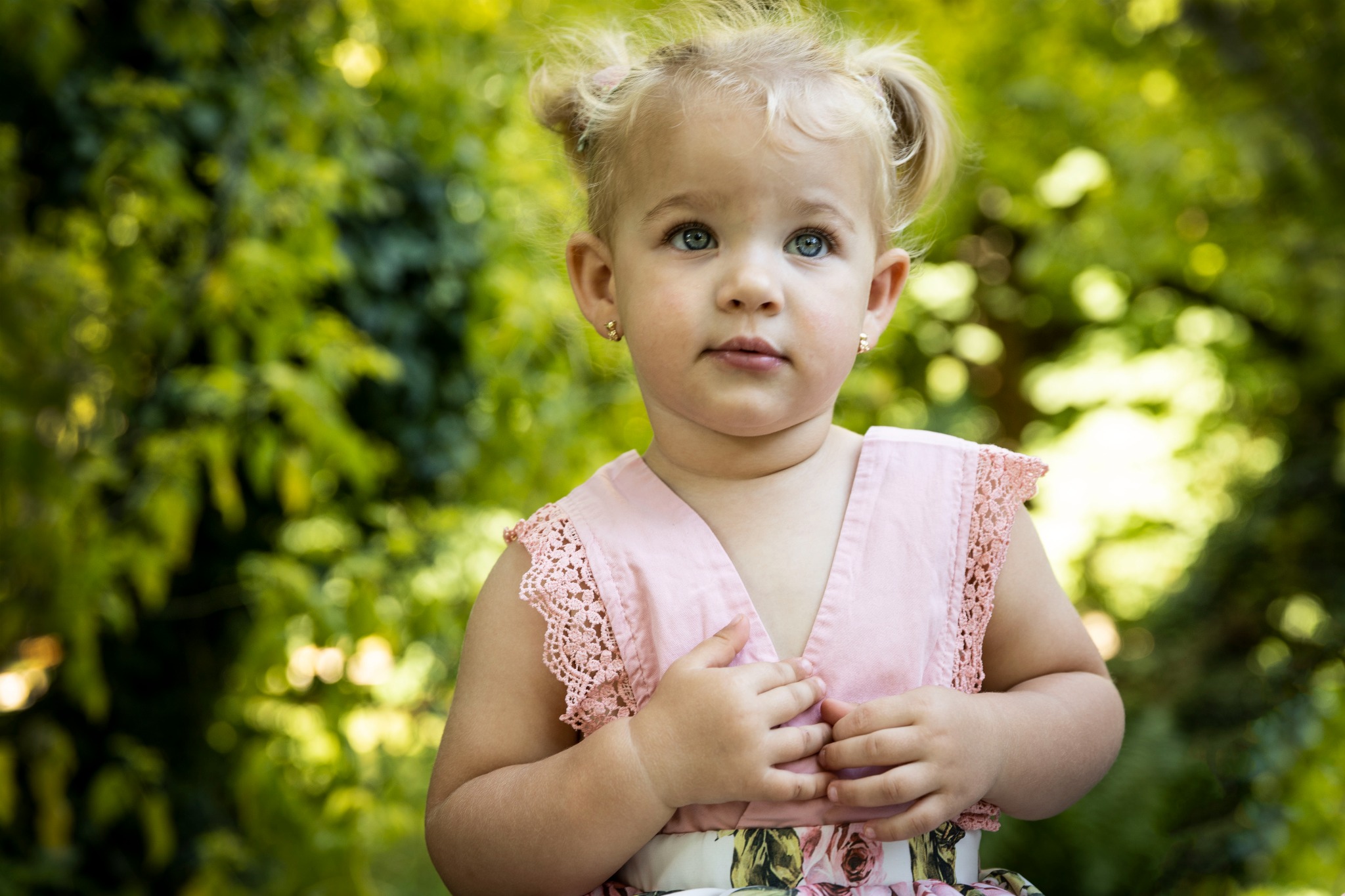 Portret uroczej, blondwłosej dziewczynki z niebieskimi oczami, w różowej sukience z koronkowymi rękawkami, na tle zieleni.