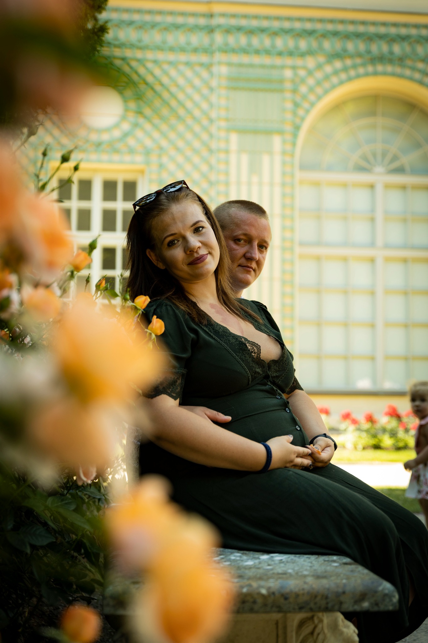 Uśmiechnięta przyszła mama w zielonej sukience, siedząca na kamiennej ławce w parku, obejmowana przez partnera, z rozkwitającymi różami na pierwszym planie i architekturą w tle.