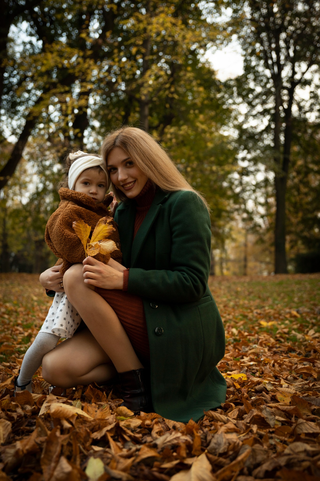 Uśmiechnięta mama w zielonym płaszczu kuca z córeczką w brązowej kurtce trzymającą żółte liście na tle jesiennego parku.