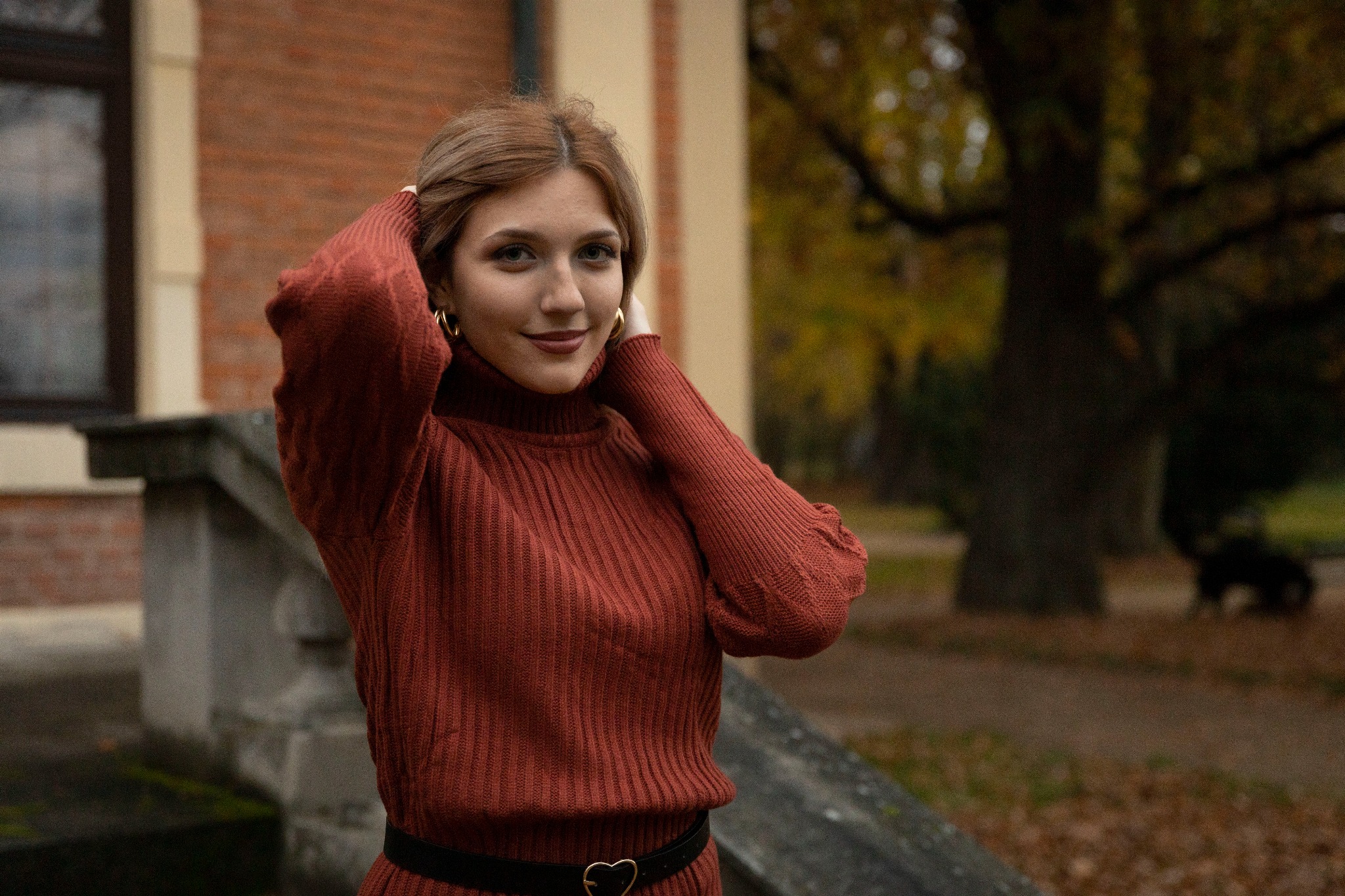 Portret kobiety w rudym swetrze z golfem i paskiem z serduszkiem, na tle ceglanego budynku i jesiennego parku.