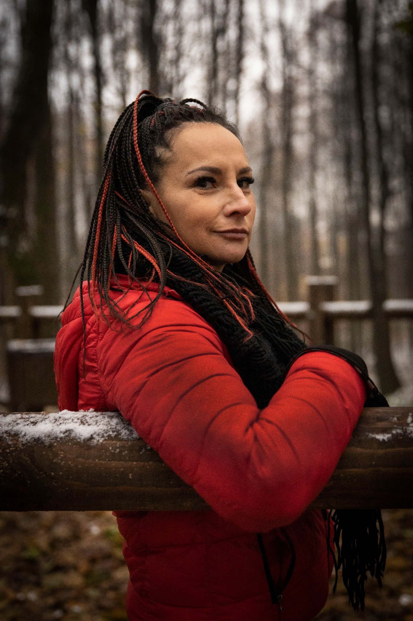 Kobieta w czerwonej kurtce opiera się o drewnianą barierkę na tle zimowego lasu, z wplecionymi czerwonymi pasemkami w ciemnych warkoczach.