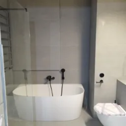 Nowoczesna, minimalistyczna łazienka w odcieniach szarości z białą, wolnostojącą wanną i toaletą z zabezpieczeniem w Piasecznie. Czarna armatura i szklana kabina prysznicowa.