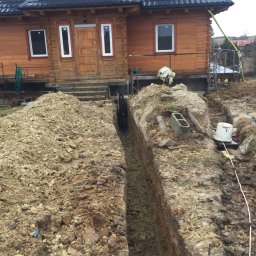 Damian Cichański - Wykopany wąski rów w ziemi na posesji z drewnianym domem w tle, prawdopodobnie pod fundament, z widocznymi narzędziami i osobą w oddali.