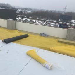 Damian Cichański - Prace izolacyjne na dachu budynku w trakcie budowy, widoczne rolki papy, żółta folia ochronna, bloczki betonowe, wełna mineralna oraz elementy mocujące na tle zabudowy jednorodzinnej i pochmurnego...