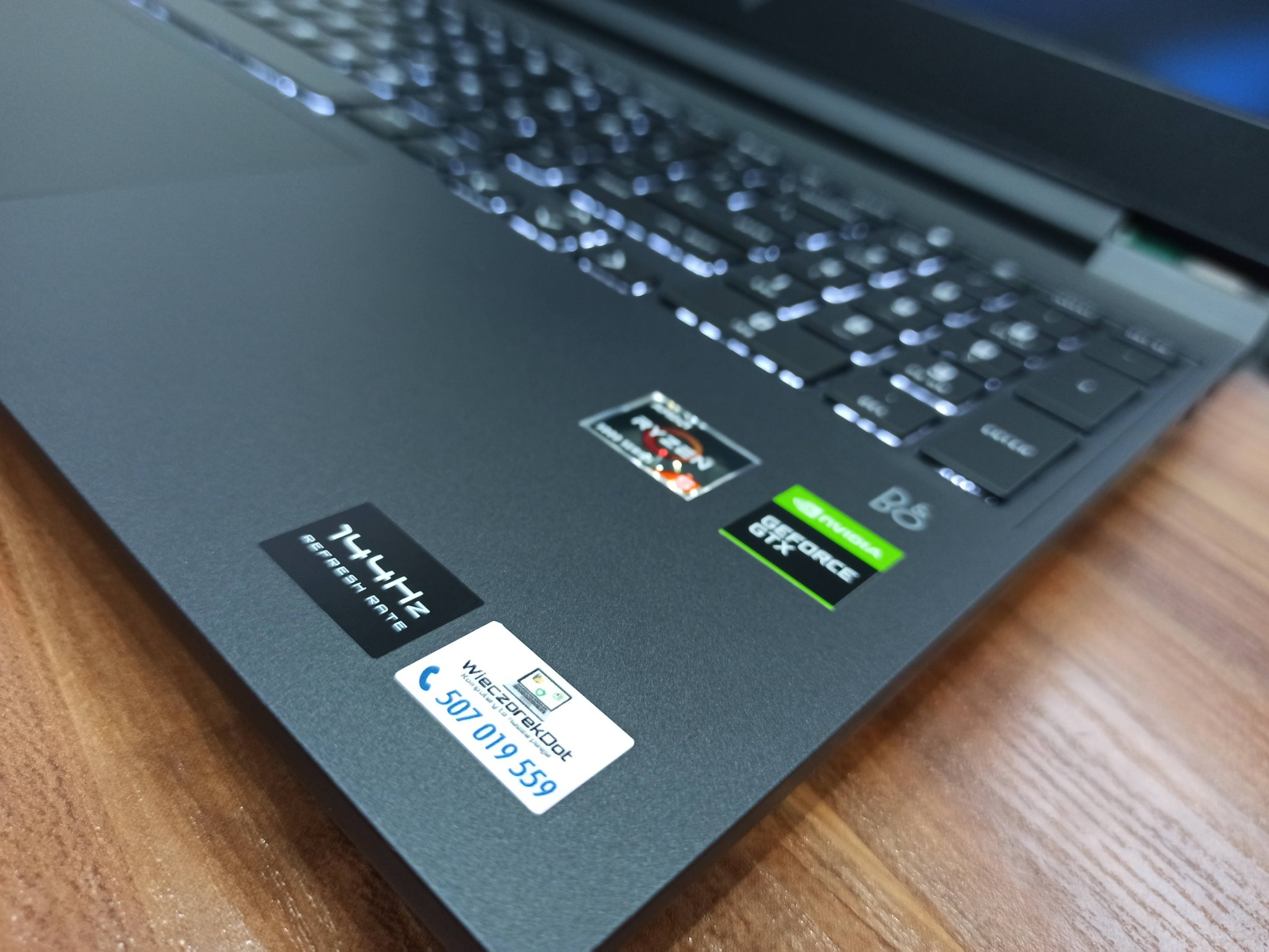 Szary laptop z podświetlaną klawiaturą, naklejkami Ryzen, Nvidia GeForce GTX, 144Hz refresh rate oraz wizytówką serwisu komputerowego WieczorekDot z numerem telefonu 507 019 559
