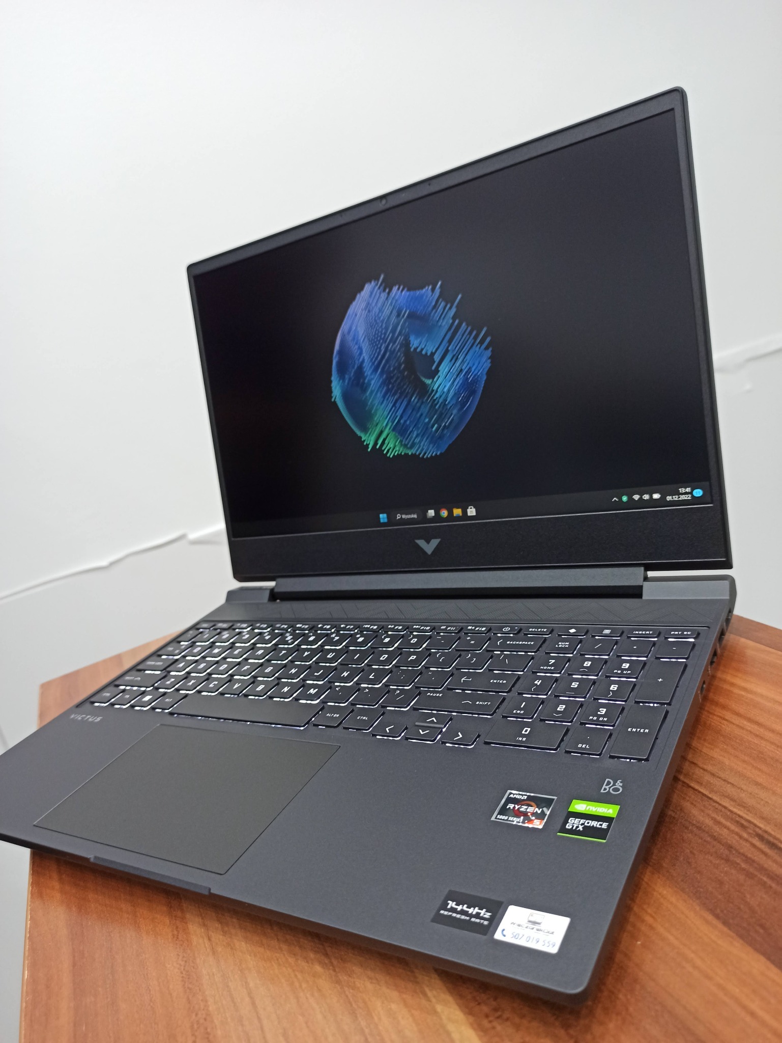 Czarny laptop Victus z podświetlaną klawiaturą, widoczny na drewnianym stole, z ekranem wyświetlającym abstrakcyjną grafikę w odcieniach niebieskiego i zielonego. Naklejki AMD Ryzen i Nvidia...