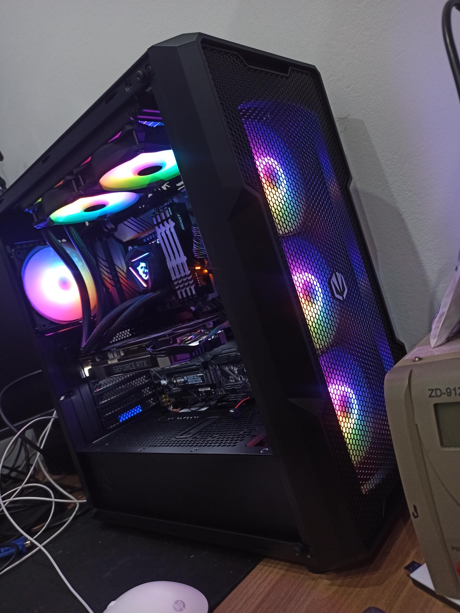 Otwarta obudowa komputera stacjonarnego z widocznymi podświetlanymi wentylatorami RGB, płytą główną MSI i kartą graficzną GeForce RTX, leżąca na biurku obok białej myszki HP i zasilacza...