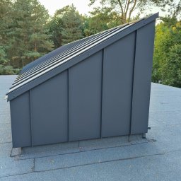 Ivan Marchenko VANBUD - Szare, metalowe obłożenie komina na dachu pokrytym papą, widoczne na tle zielonych drzew i jasnego nieba. Detal obróbki blacharskiej.