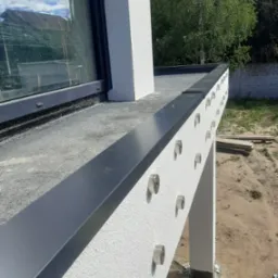 Nowoczesny dach płaski z czarnym wykończeniem krawędzi i mocowaniami punktowymi na ścianie attyki, widok z zewnątrz w słoneczny dzień.