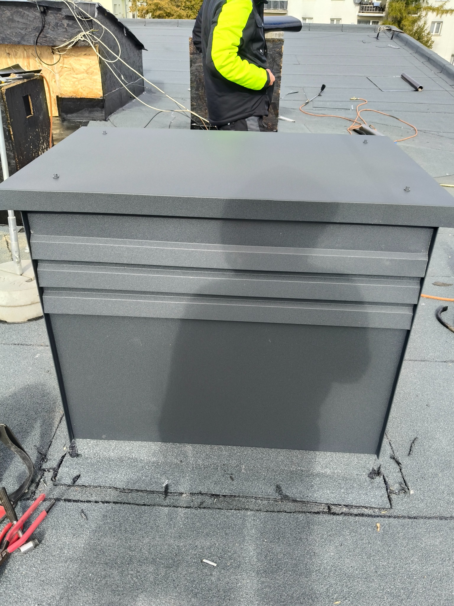 Szara, metalowa obudowa komina na płaskim dachu z papy, widoczne narzędzia dekarskie i fragment sylwetki dekarza w odblaskowej kurtce. Detal wykończenia dachu.