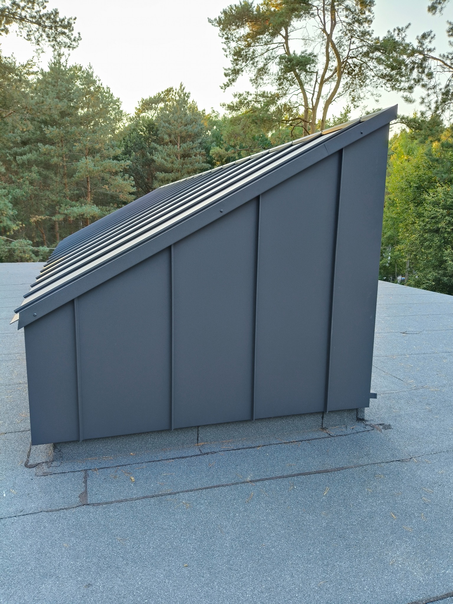 Szare, metalowe obłożenie komina na dachu pokrytym papą, widoczne na tle zielonych drzew i jasnego nieba. Detal obróbki blacharskiej.