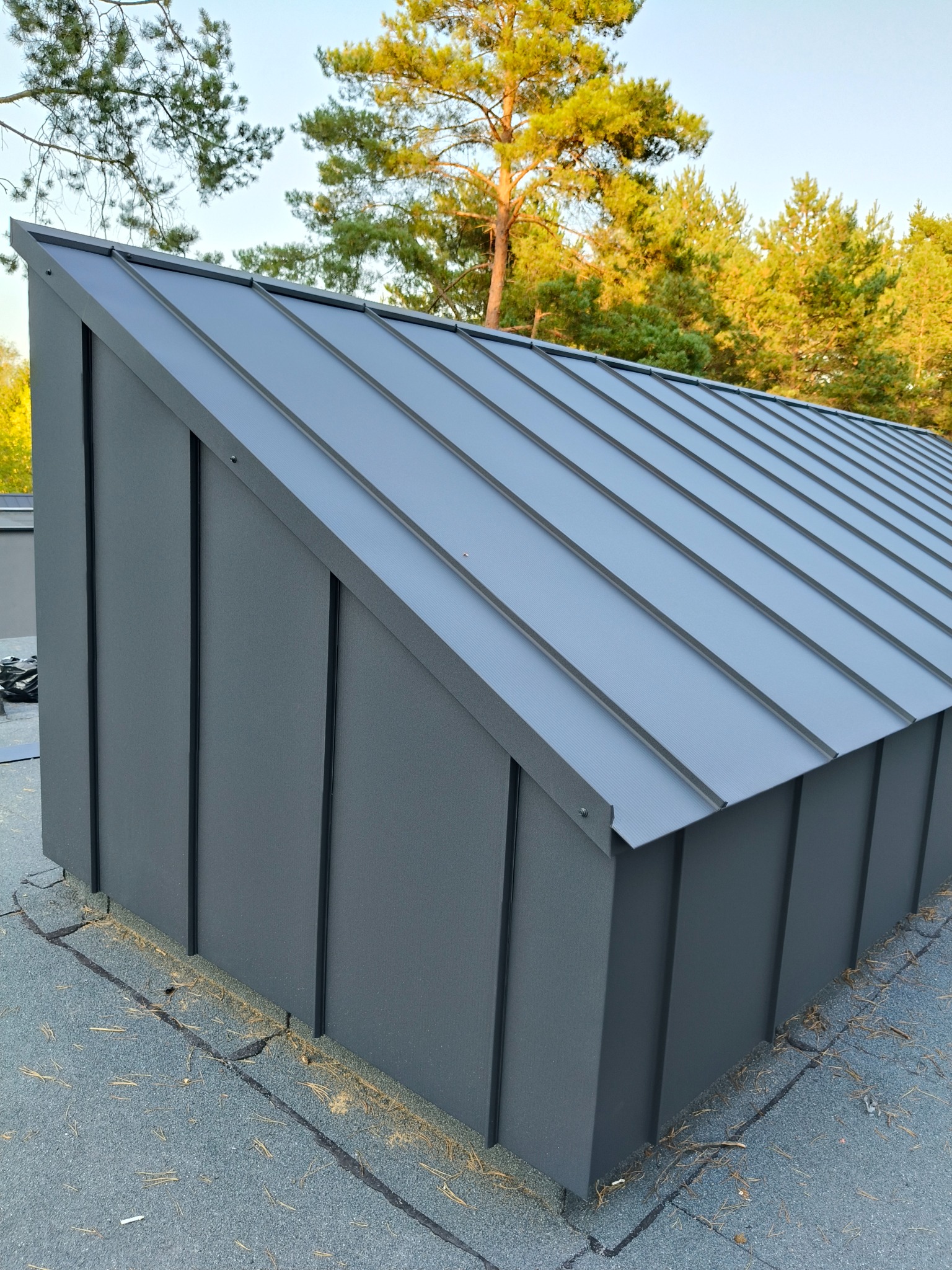 Nowoczesne, grafitowe krycie dachu z blachy na budynku z widocznym tłem drzew. Estetyczne wykończenie detali i precyzyjne łączenia blacharskie.