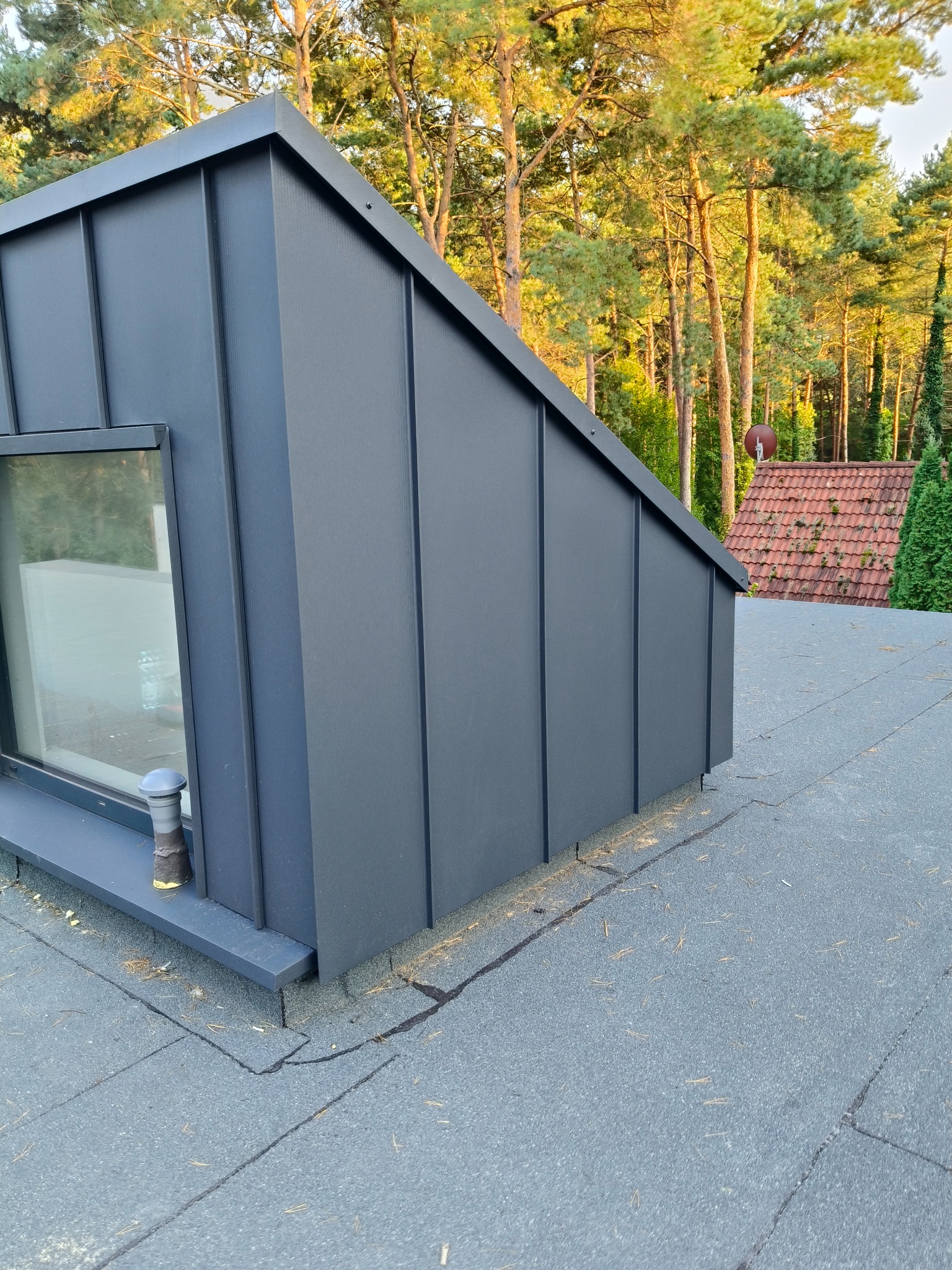 Szary lukarna z oknem na dachu pokrytym papą, widok z góry. W tle drzewa i dach innego budynku.