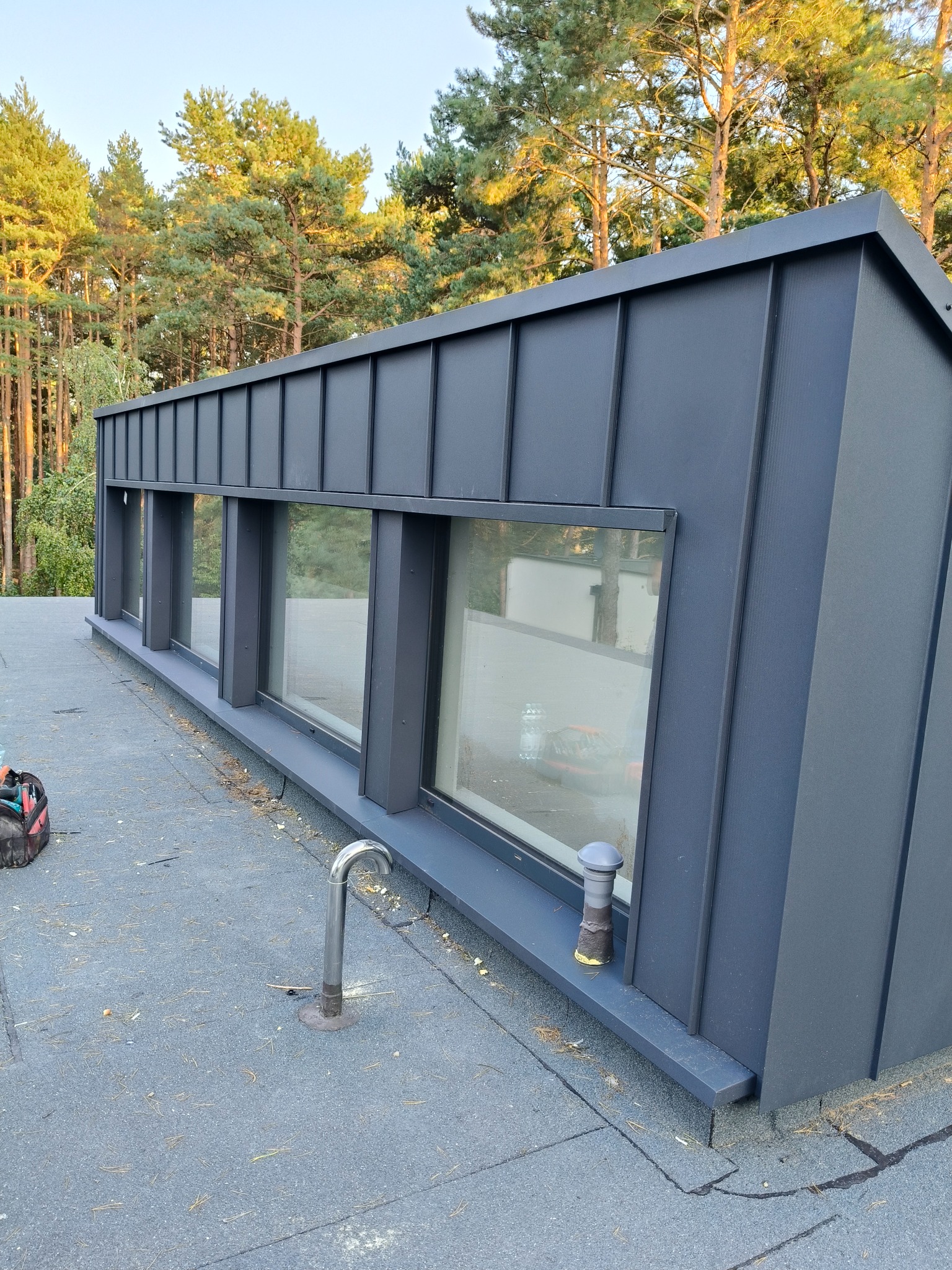 Nowoczesna, grafitowa obudowa lukarny z trzema oknami, wykończona blachą na dachu pokrytym papą. W tle widoczne drzewa iglaste.