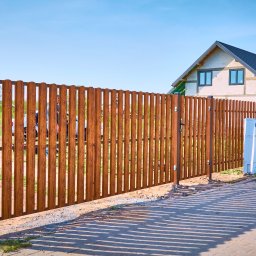 Steel Mark - Nowoczesne, drewniane ogrodzenie panelowe z pionowych desek w kolorze brązowym, z widoczną furtką i słupkami, na tle domu w stanie surowym i błękitnego nieba. Długie cienie rzucane przez ogrodzenie...