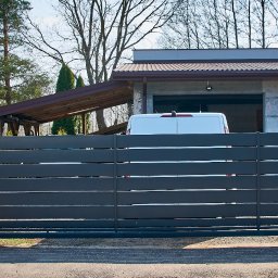 Steel Mark - Nowoczesna, przesuwna brama z poziomych, szarych profili, osadzona na słupkach z dekoracyjnych, brązowo-beżowych bloczków betonowych. W tle widoczny budynek mieszkalny z białym vanem zaparkowanym...