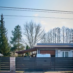 Steel Mark - Nowoczesna, pozioma brama wjazdowa z szarych paneli metalowych, z jasnymi słupkami z imitacji kamienia, na tle domu z garażem i podjazdem, w słoneczny dzień.