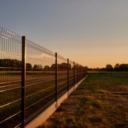 Steel Mark - Długi, brązowy płot panelowy z podmurówką betonową oddzielający pole od łąki, sfotografowany pod kątem podczas zachodu słońca.