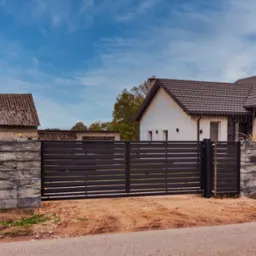 Nowoczesne, poziome przęsła w kolorze antracytowym tworzą ogrodzenie posesji z jasnym domem i starą stodołą w tle. Słupki ogrodzeniowe z szarej kostki betonowej. Skrzynka pocztowa z numerem 50.