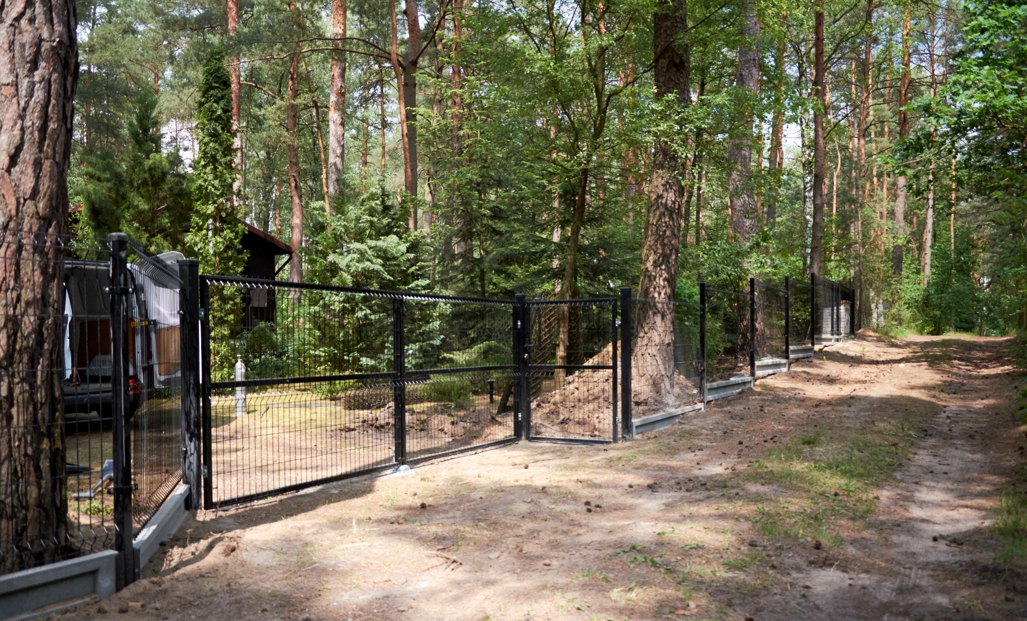Czarny panelowy system ogrodzeniowy z furtką, zamontowany na betonowej podmurówce, otoczony leśnym krajobrazem z wysokimi drzewami i piaszczystą ścieżką, z widocznym busem w tle.