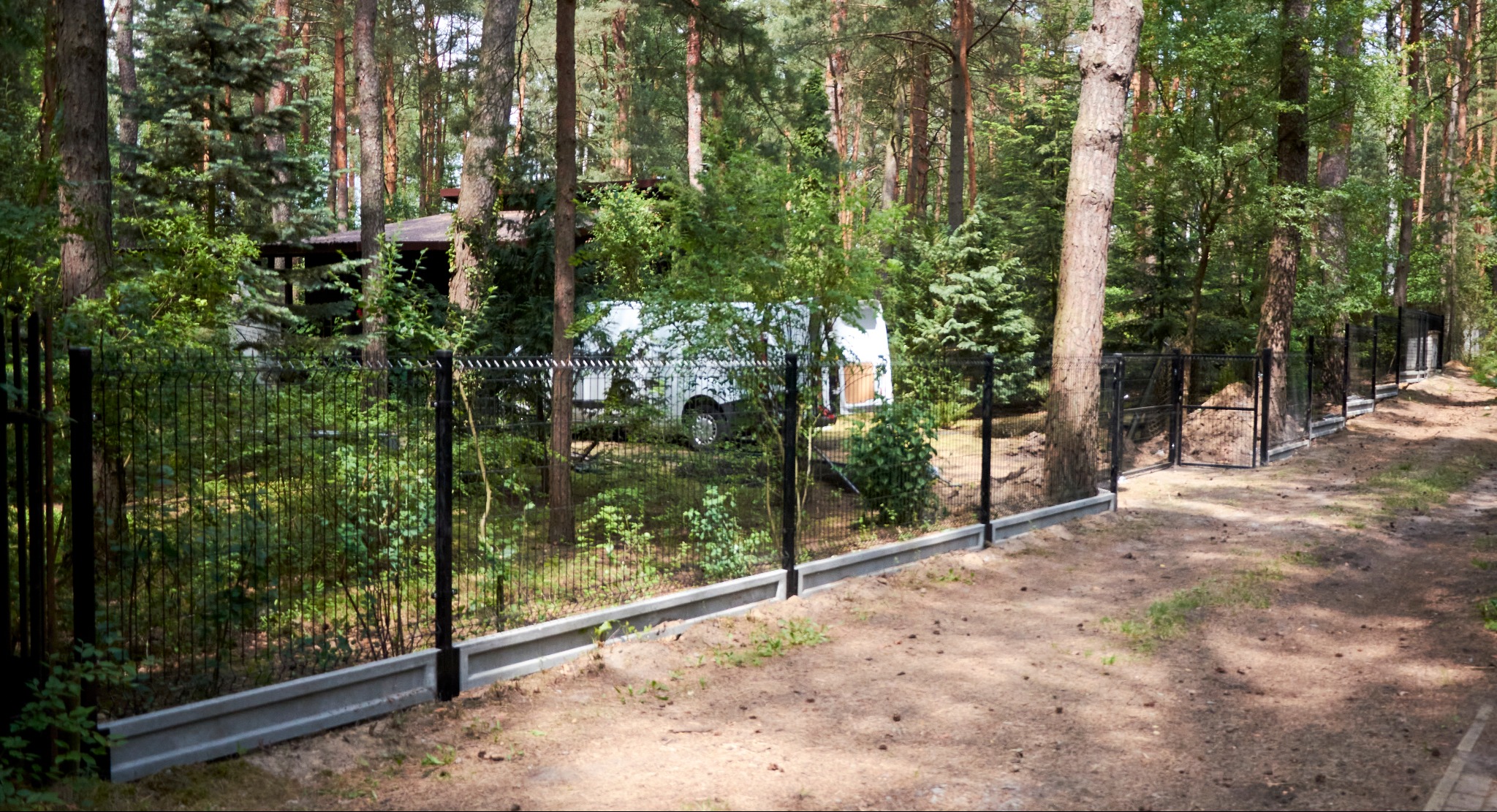 Czarny panelowy płot z podmurówką betonową w leśnej scenerii, z bramką i białym busem w tle.