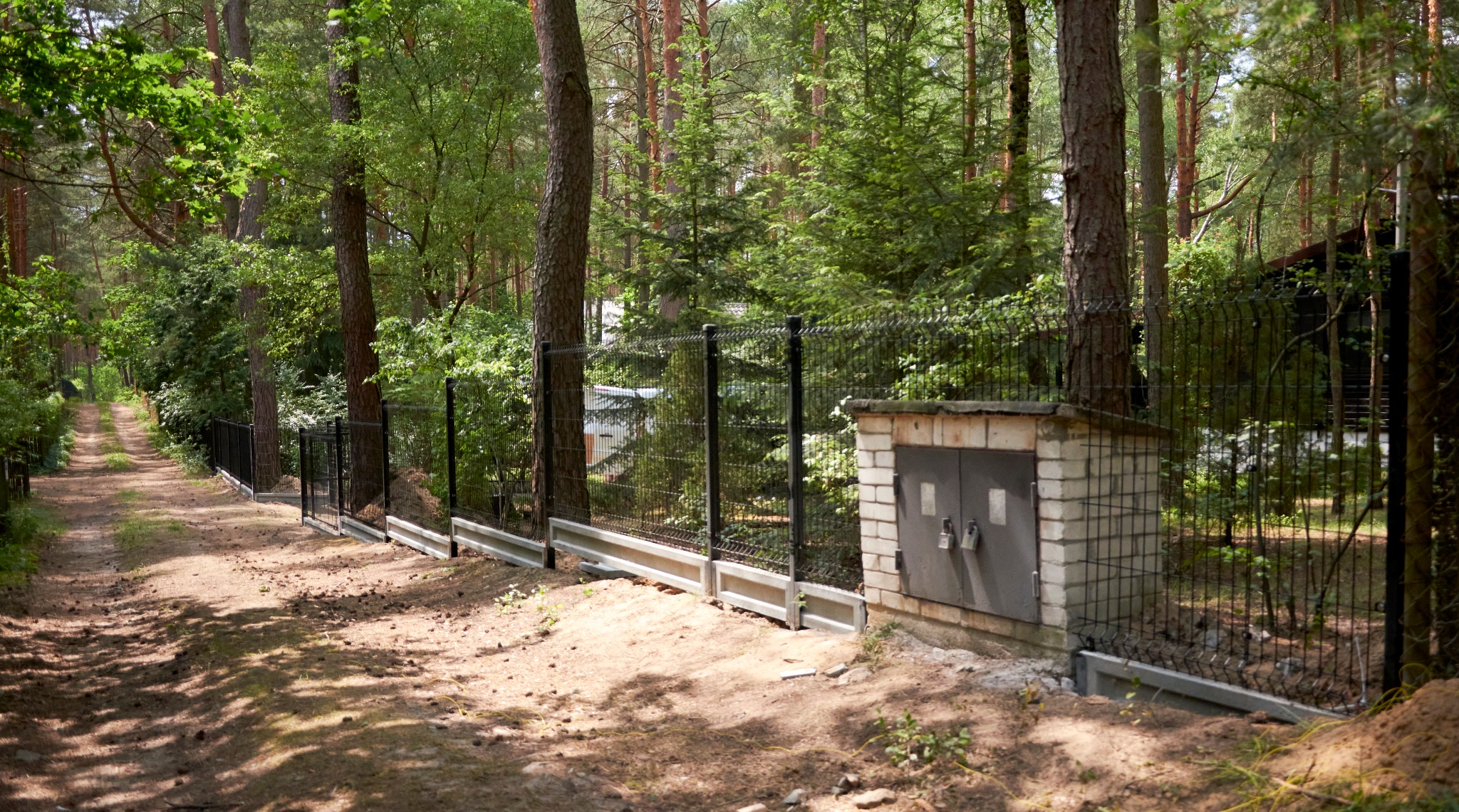 Czarny panelowy system ogrodzeniowy z podmurówką betonową biegnący wzdłuż leśnej drogi, z widoczną skrzynką elektryczną z cegły tuż za ogrodzeniem.